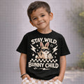 Olive-skinned young boy with short brown hair wearing a black “Stay Wild Bunny Child” Easter graphic T-shirt with cool bunny design and beige check shorts.