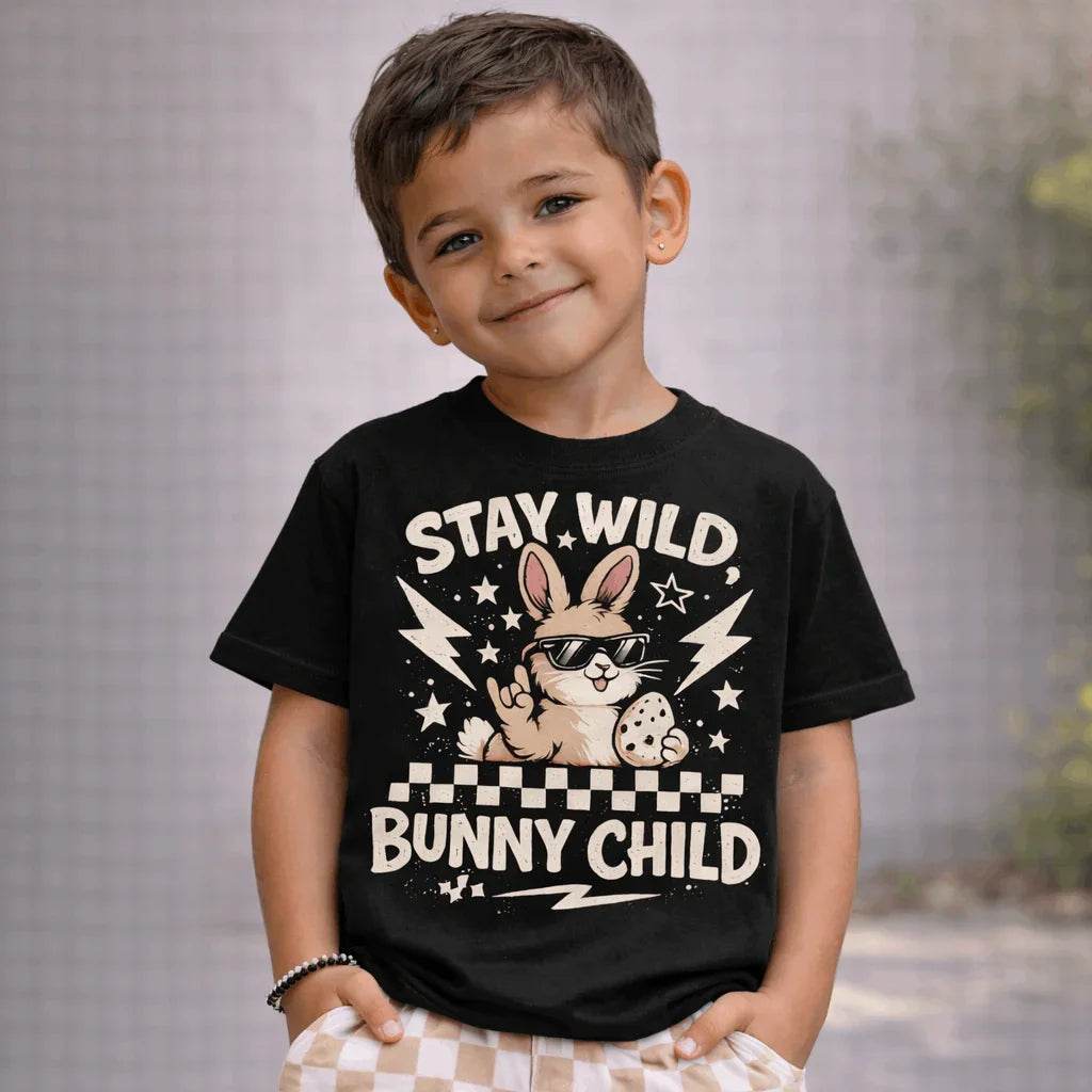 Olive-skinned young boy with short brown hair wearing a black “Stay Wild Bunny Child” Easter graphic T-shirt with cool bunny design and beige check shorts.