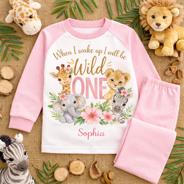Personalised wild one birthday pyjamas for girls featuring safari animals and floral design with the text “When I wake up I will be Wild One” and custom name on white and pink children’s pyjama set flat lay.